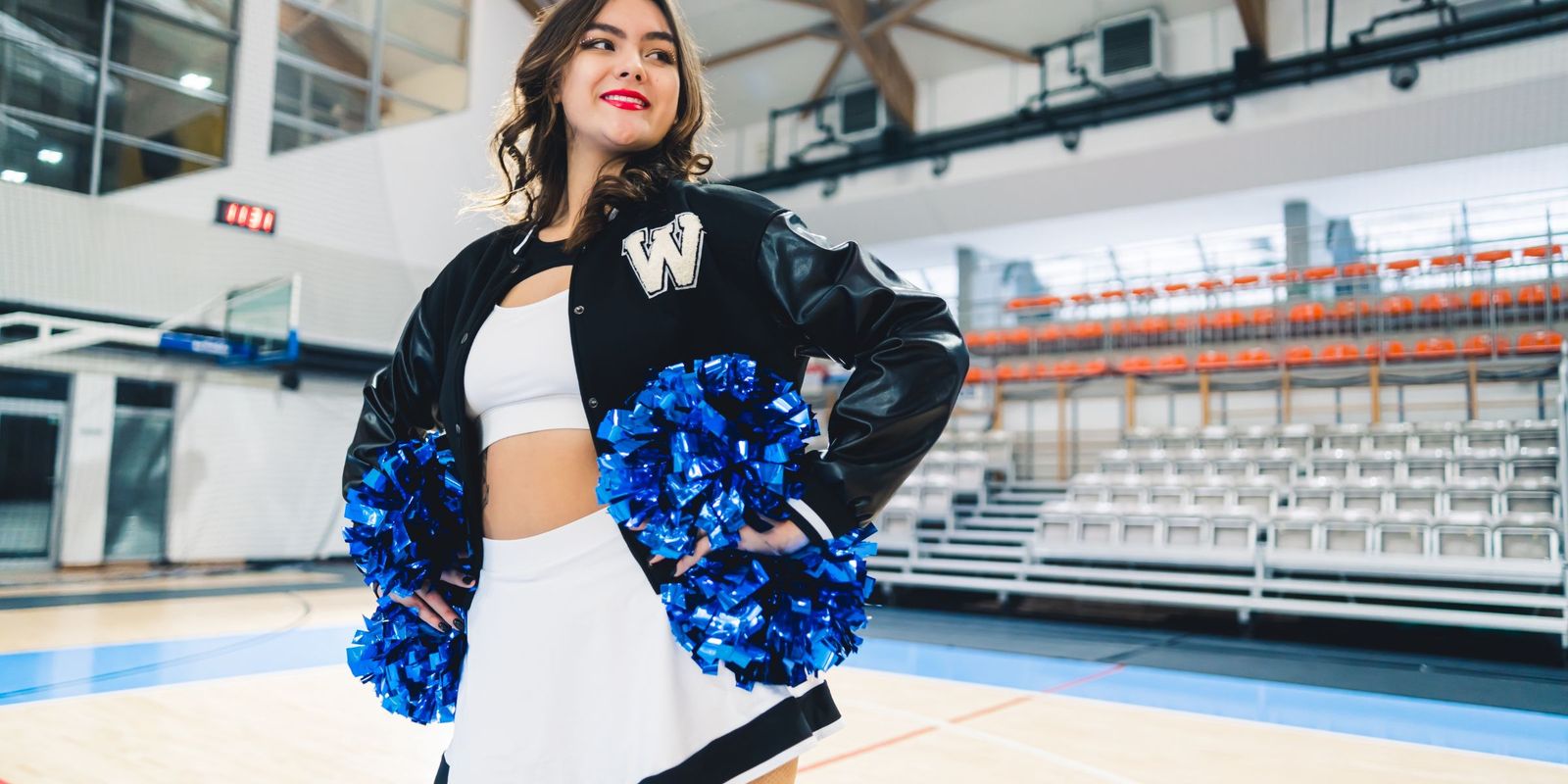 A confident cheerleader standing in an empty gym, blue pom-poms in hand | Source: Shutterstock
