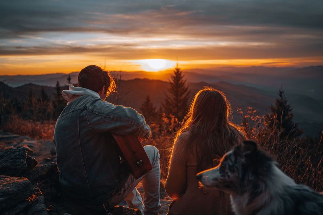Silhouetted couple with dog capturing tranquil mountain sunset, creating a serene getaway moment.