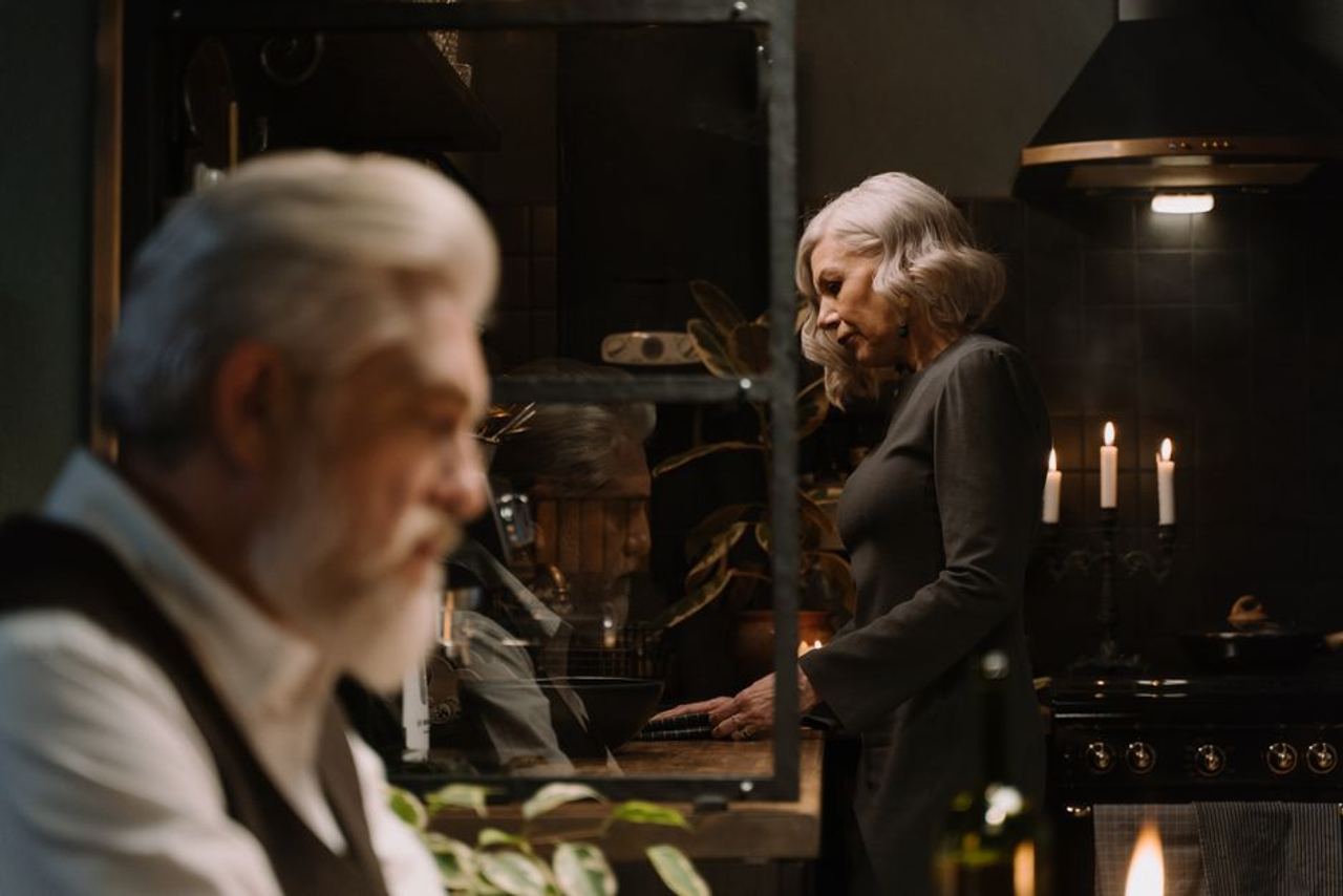 A distinguished elderly couple in a cozy, warm kitchen setting with candles and plants.