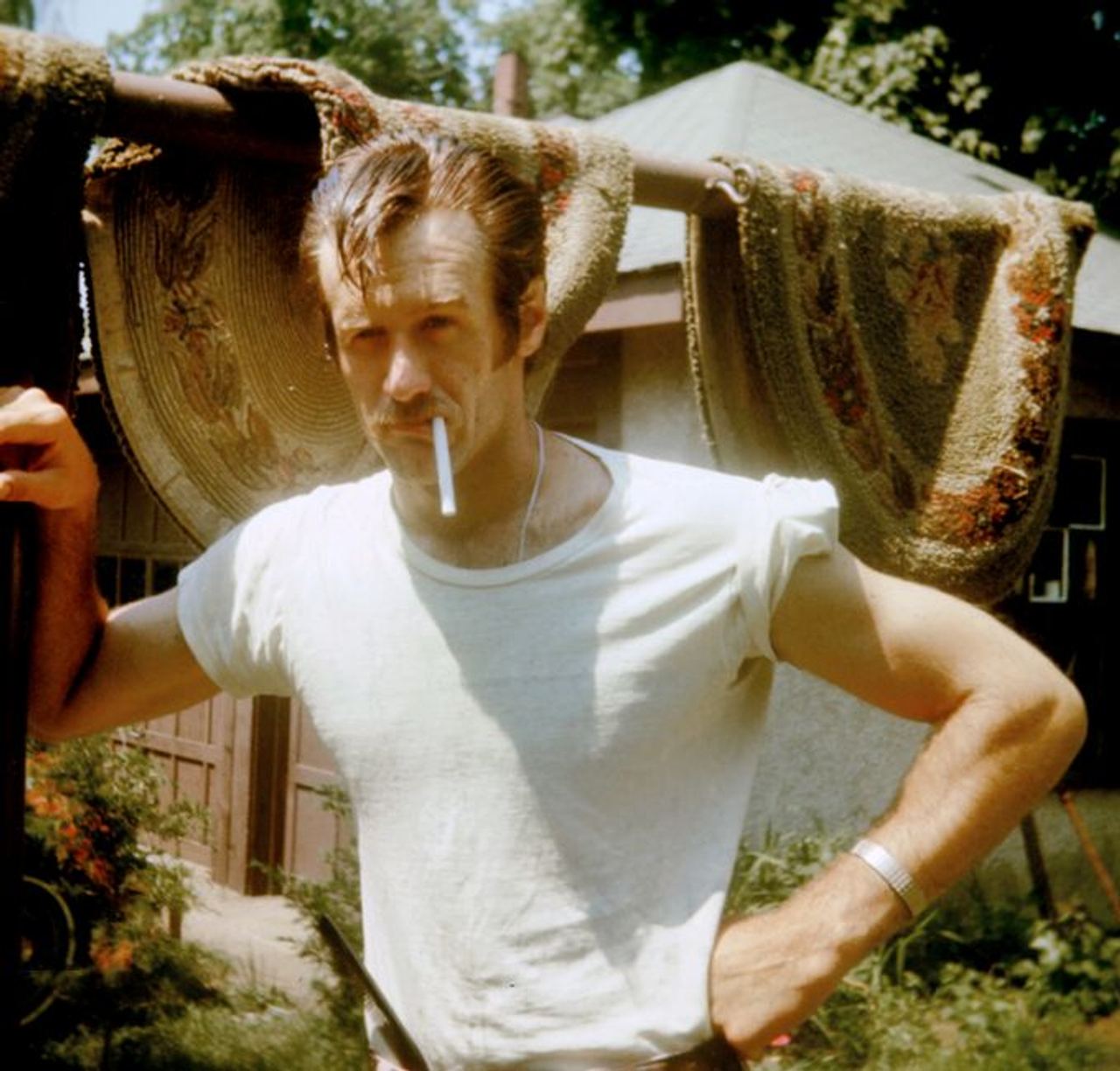 A stylish man in casual wear poses outdoors with a cigarette, showcasing a relaxed summer vibe.