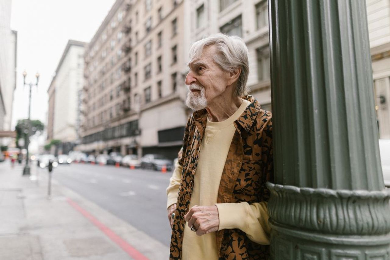 A senior man with a beard leaning on a pole on an urban street, standing in side view.