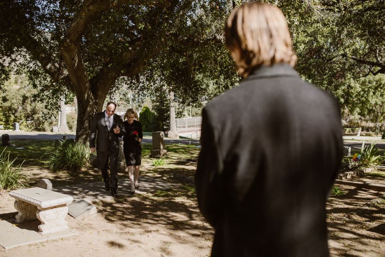 A couple walks through a cemetery mournfully, surrounded by gravestones and nature.