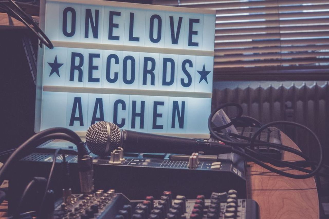 A warm-toned studio featuring a microphone, audio mixer, and lightbox signage reading 'One Love Records Aachen'.